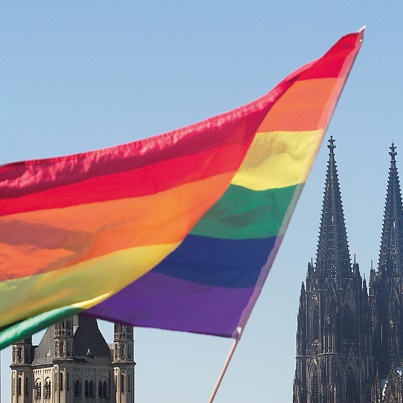Eine Regenbogenflagge weht im Wind. Im Hintergrund befindet sich der Kölner Dom.