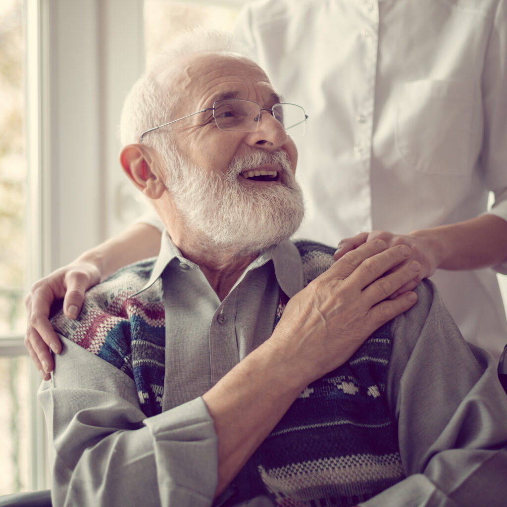 Ein älterer Mann sitzt lachend und die Hand seiner Krankenschwester haltend im Rollstuhl